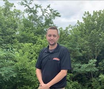Male employee outdoors wearing black SERVPRO shirt standing in front of trees